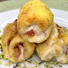 Tre involtini di pollo dorati e ripieni di formaggio filante e prosciutto, adagiati su un letto di pistacchi tritati.