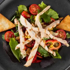 Deliziosa insalata di pollo fredda con pomodorini, mais, peperoni e crostini croccanti su un piatto scuro.