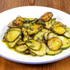 Piatto di zucchine alla poverella, fette sottili e dorate condite con olio e erbe aromatiche, su un piatto bianco con bordo blu.