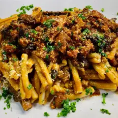 Un piatto di pasta alla norma fumante, con melanzane fritte, pomodoro e ricotta salata grattugiata, guarnito con prezzemolo fresco.
