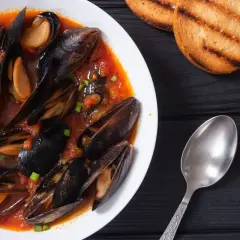 Un invitante piatto di zuppa di cozze in un ricco sugo di pomodoro, guarnito con erba cipollina, accompagnato da crostini tostati e un cucchiaio.
