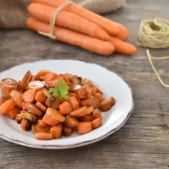 Carote in padella, dolci e caramellate, guarnite con prezzemolo fresco su un piatto bianco. Sullo sfondo, carote fresche legate con spago.