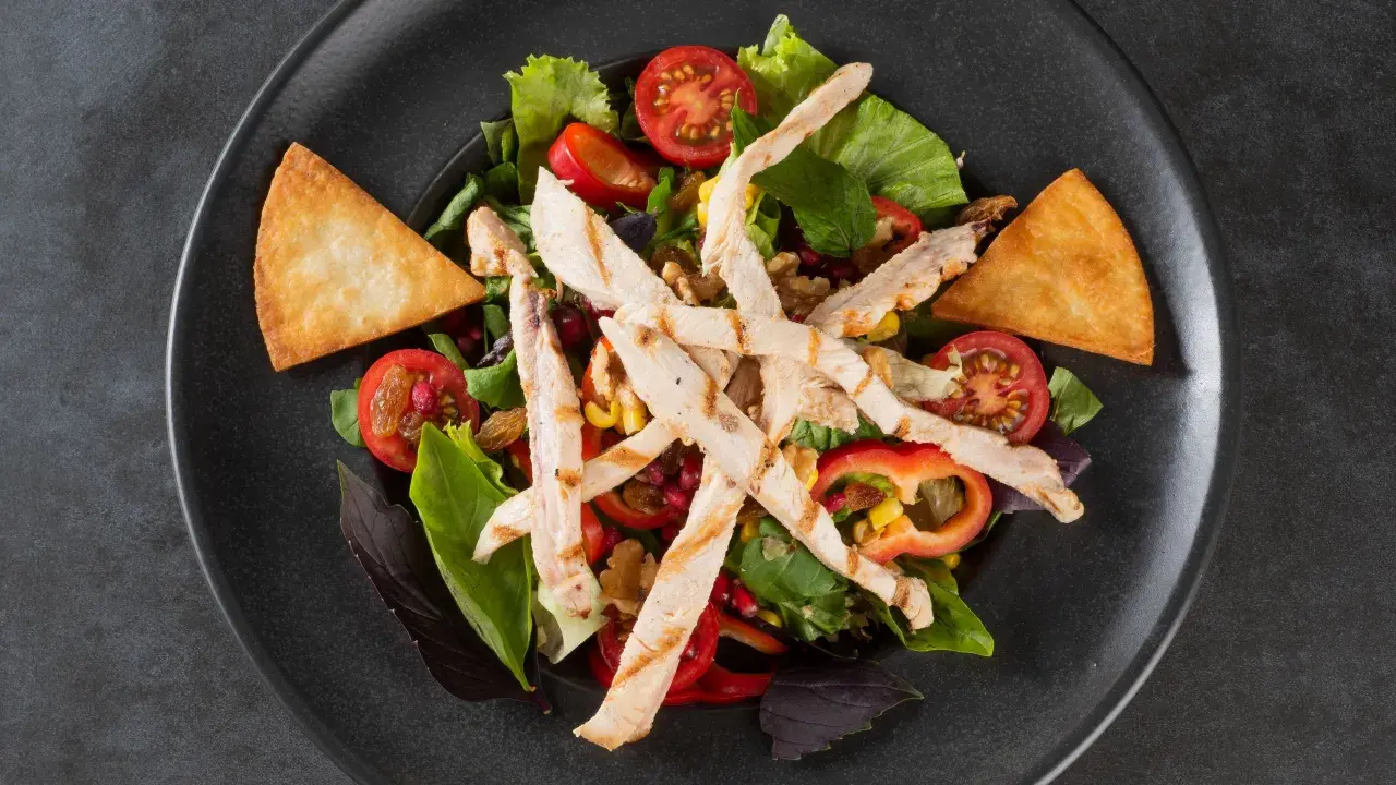 Deliziosa insalata di pollo fredda con pomodorini, mais, peperoni e crostini croccanti su un piatto scuro.