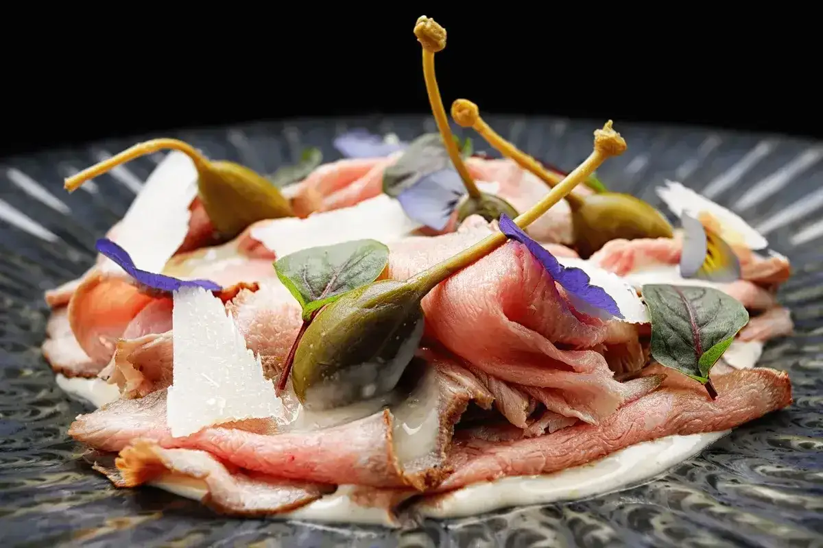 Vitello tonnato veloce: fette sottili di arrosto di vitello con salsa tonnata, capperi, scaglie di parmigiano e fiori eduli su piatto scuro.