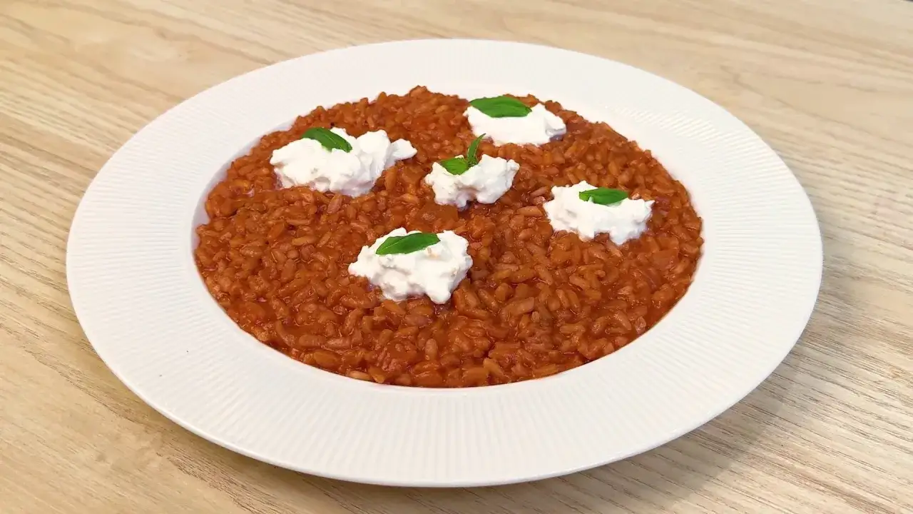 Un cremoso risotto al pomodoro, arricchito da ciuffi di ricotta fresca e foglie di basilico, servito su un piatto bianco.
