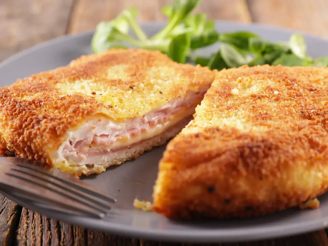 Cotoletta alla valdostana dorata e croccante, ripiena di prosciutto e formaggio filante, servita su un piatto grigio con un contorno di insalata verde.
